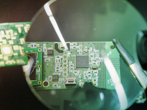 Unter einer Lupe befindet sich ein grünes PCB-Board mit kleinen elektronischen Bauteilen.