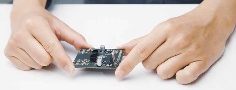 Ein grünes PCB-Board mit Bauteilen wird von zwei Händen auf einem weißen Tisch gehalten.
