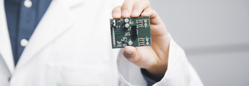 Startseite_Header Ein Mensch in einem weißen Kittel hält mit einer Hand ein grünes PCB-Board mit Bauteilen.