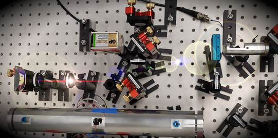 Viele optische Komponenten wie Spiegel, Linsen, Laser und ein großes Rohr aus Metall auf einem optischen Tisch mit regelmäßigen Bohrungen.