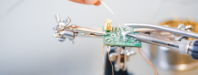 Über einem grünen PCB-Board mit Bauteilen wird ein Lötkolben gehalten. An die Spitze des Lotkolbens halten zwei Finger einen Lötzinn, der in eine weiße Wolke verdampft.