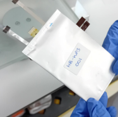 Zwei Hände in blauen Handschuhen halten eine handgroße Batteriezelle. Aus der Zelle kommen 5 Streifenleiter.